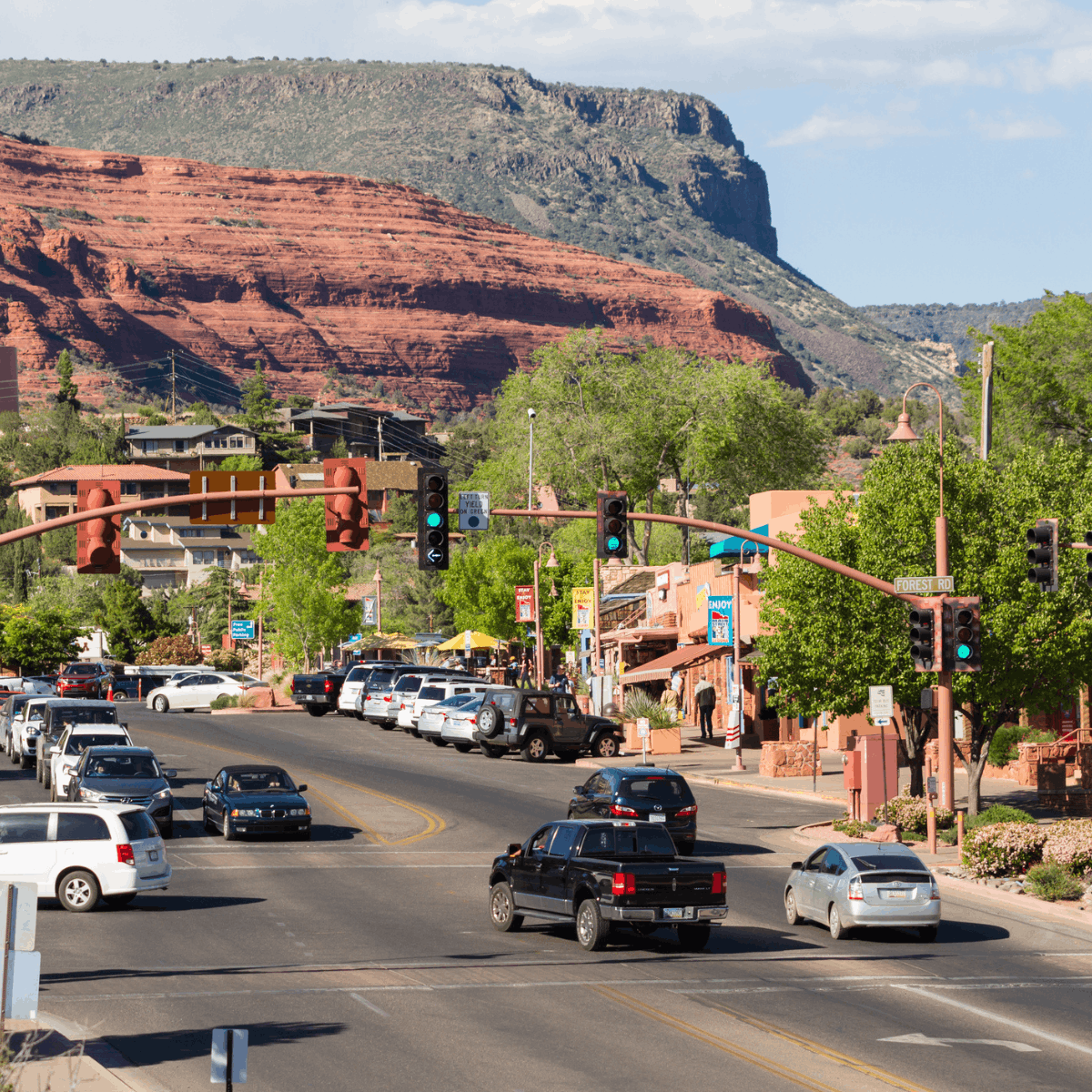 Busy desert town street with cars, storefronts, and traffic lights against red rock cliffs in the background.
