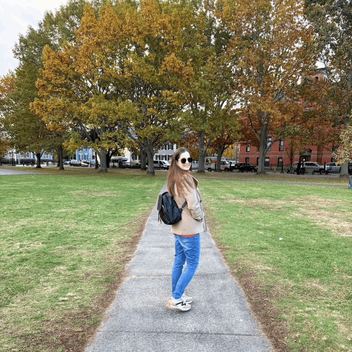 Woman walking along a park path with autumn trees in the background, wearing a beige coat, blue jeans, sneakers, sunglasses, and a black backpack.