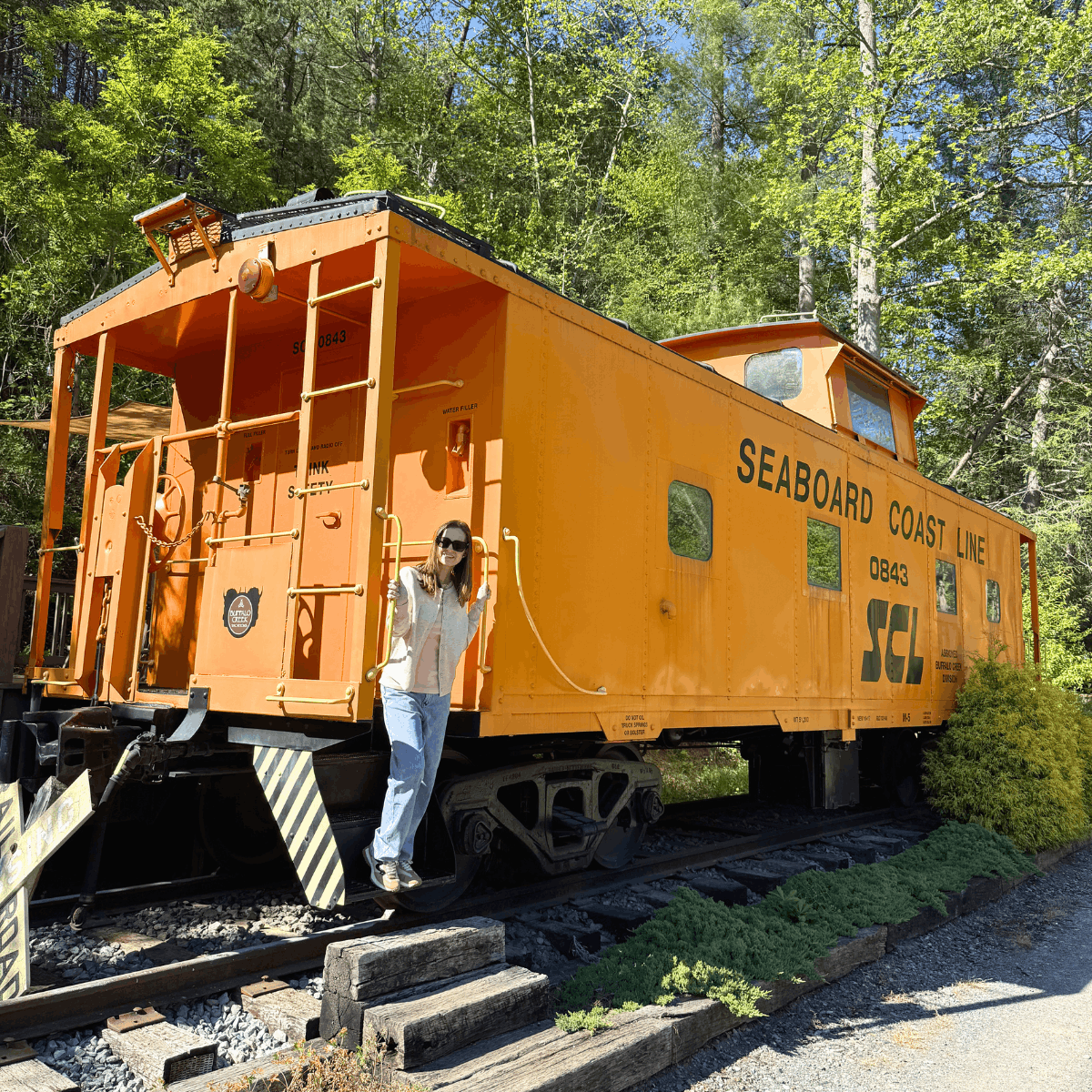Bright orange Seaboard Coast Line maintenance railcar on tracks, with a woman standing at the doorway in a wooded area.